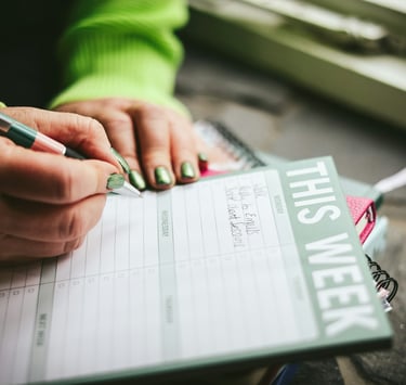 A person writing in a green weekly planner pad to organize their schedule and tasks.