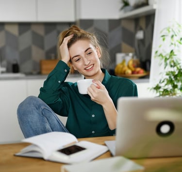 Smiling woman drinking coffee while working on a laptop in a modern kitchen home office.