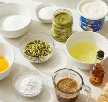 Baking ingredients for pistachio tiramisu featuring mascarpone, ladyfingers, espresso, and pistachio cream on a counter.