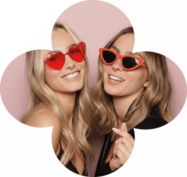 Two smiling women wearing stylish red heart-shaped and cat-eye sunglasses against a pink background.