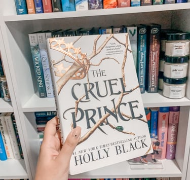 Mariah is holding up a hardcover copy of The Cruel Prince by Holly Black in front of her shelves.