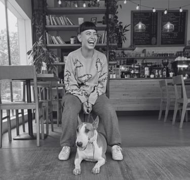 Mujer riendo sentada junto a su Bull Terrier Inglés en una cafetería moderna.