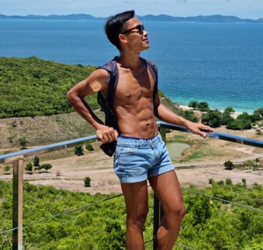 Man wearing blue shorts and no top standing on a viewpoint with sea and an island in the background.