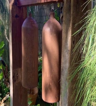 Rustic iron wind chimes hanging from a weathered wooden frame in a lush garden setting.