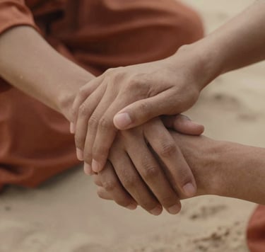 A close-up, authentic shot of two hands gently clasped together, capturing a moment of genuine human connection. The scene is bathed in a warm, inviting glow with terracotta-colored fabrics in the background and soft sand skin tones.