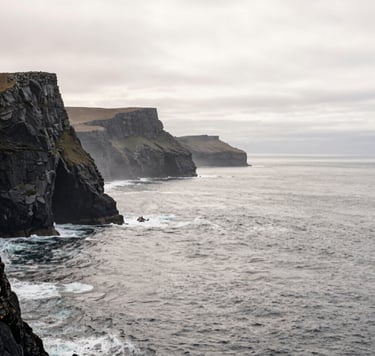 A minimalist, wide-angle landscape photograph of a rugged coastline in a Global / Western setting. The scene features Charcoal Grey cliffs and a Soft Off-White mist rolling over the Atlantic ocean, captured with a storytelling, adventurous aesthetic.