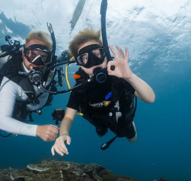 A scuba diving instructor and child giving an okay sign underwater near a coral reef.