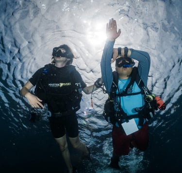 Two scuba divers performing a safety stop underwater with sunlight shimmering through the surface.