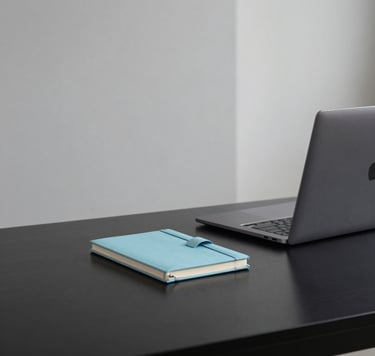 A minimalist studio desk in a South American / Brazilian office, featuring a sleek black surface, a silver laptop, and a small baby blue decorative notebook. The lighting is soft and natural, emphasizing clean dark gray and light gray lines in a professional setting.