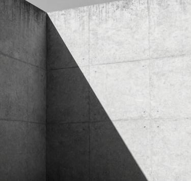 A high-contrast black and white architectural photograph of a minimalist concrete structure in a North American setting. Sharp geometric shadows fall across a light gray textured wall, emphasizing clean lines and sophisticated architectural forms. Editorial style.