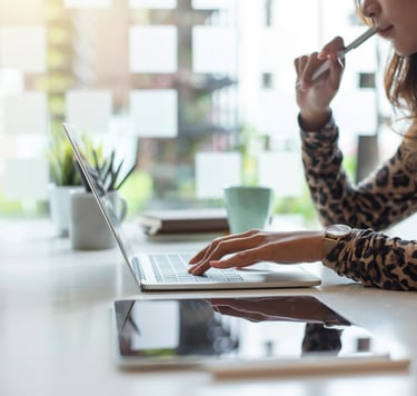 Professional woman holding a pen near her mouth, working on a laptop on a desk with a digital tablet, plant, and coffee cup.