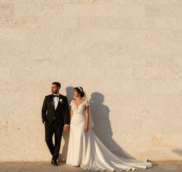 Professional outdoor wedding photography in Bodrum at golden hour, a bride and groom standing against a clean white stone wall, soft mustard yellow sunlight, minimal and modern composition, Middle Eastern / Turkish wedding details, warm and professional atmosphere.