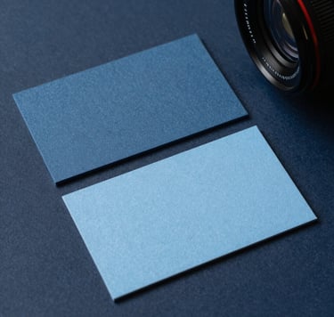 A close-up photograph of high-quality, textured business card samples in Muted Slate Blue and Soft Sky Blue on a clean Dark Navy surface. Professional studio lighting in a modern International / Western design office, showcasing a clean and sophisticated aesthetic.