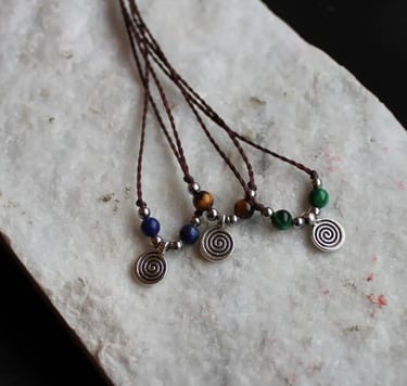 Drei Ketten mit braunem Garn und einem silberfarbenen Spiralenanhänger. Jede Kette hat andere Perlen: blaue, grüne oder braun