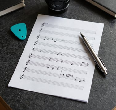 A flat lay photography shot of music notation paper and a professional silver pen on a dark charcoal desk. A bright turquoise plectrum sits nearby. The scene is set in a sunny, International / English-speaking home office environment.