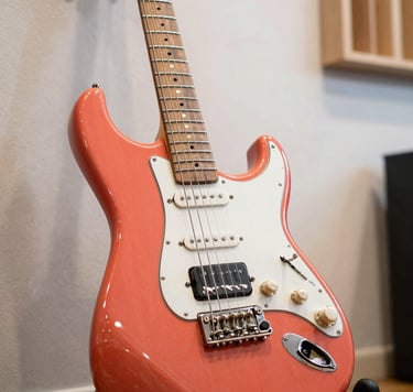 A close-up photograph of a vibrant coral electric guitar resting against a soft off-white wall in a bright, modern International / English-speaking music studio. The lighting is energetic and fresh, highlighting the wood grain and metallic hardware.