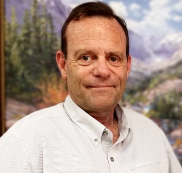 A man with dark hair, wearing a light-colored shirt. The background is a blurred landscape.