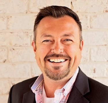 Picture of a man with brown hair and goatee, smiling. He's wearing a bright shirt and a dark suit.