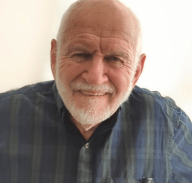 A white with a short trimmer white beard, wearing a dark patterned shirt.