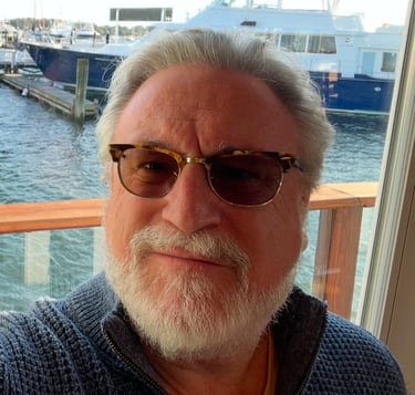 a man with a beard and sunglasses on a pier with water and boats behind him
