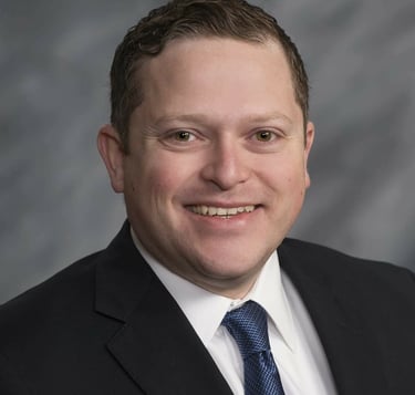 Professional headshot of a smiling man in a black suit and blue tie against a grey background.