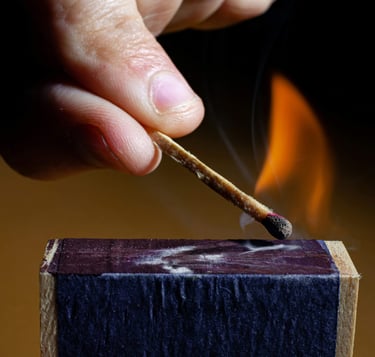 A person striking a wooden match against a matchbox, creating a bright orange flame and smoke.