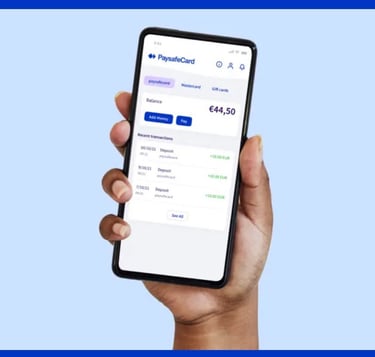 A hand holding a smartphone displaying the paysafecard app with balance and recent transactions.