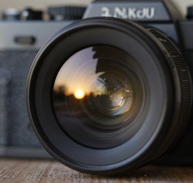 A close-up, artistic shot of a vintage camera lens reflecting a sunset scene. The glass of the lens catches glints of #C4B29A and #F5F0E1, while the body of the camera is a deep #262626. The focus is sharp on the reflections, capturing the 'moment' within the gear itself. The style is premium, sophisticated, and rich with texture.