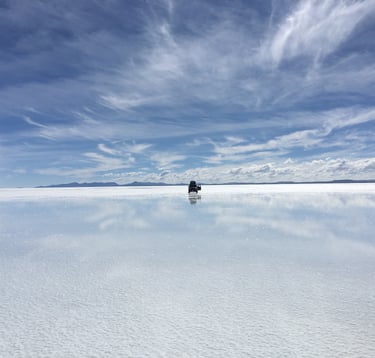 Panorama du Salar d’Uyuni reflétant le ciel bleu avec un 4x4 isolé avançant sur l’eau argentée sous des nuages légers