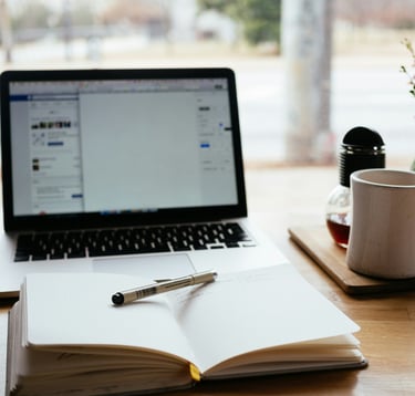 image shows an empty notebook with a pen and a laptop in the background