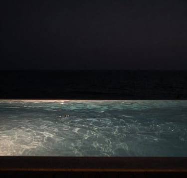 A high-contrast, artistic photography of a tranquil infinity pool meeting the dark ocean horizon at night. The water surface is like glass, reflecting a few pinpricks of soft light. Moody, cinematic atmosphere in shades of black and muted grey. North American / International context.