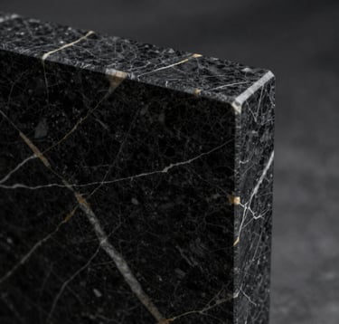 A macro photography shot of premium black marble with subtle white and gold veining. The texture is polished and reflective, capturing a sharp architectural corner in the frame. Editorial lighting with deep shadows. North American / International style.