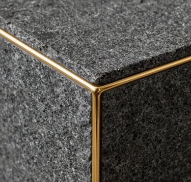 A close-up photograph of raw, dark grey textured stone meeting a thin, polished gold metal inlay. The focus is sharp on the transition between materials, emphasizing craftsmanship and luxury textures. North American / International.