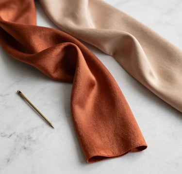 An editorial still life featuring high-quality silk threads in terracotta and warm beige, draped elegantly over a minimalist marble surface. A vintage brass needle sits nearby. The style is sophisticated and clean. North American / US luxury design context.