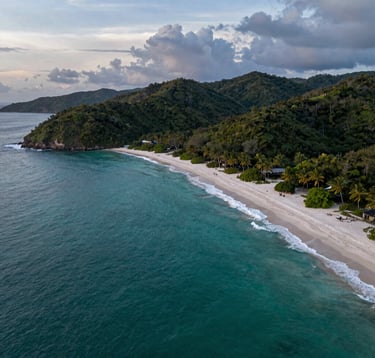 Dramatic aerial photography of the Guanacaste coastline at sunset, featuring the transition from turquoise ocean waters to the white sand beaches of Playa Flamingo and lush green hills. The composition highlights the geographical exclusivity of the region. Soft steel blue and light grey tones in the sky, professional landscape photography.