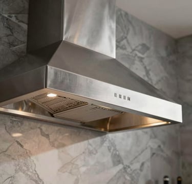 Close-up of a wall-mounted stainless steel hood in a luxury Brazilian kitchen. The finish is brushed inox with champagne lighting accents. High-end materials like marble and graphite are visible in the background.