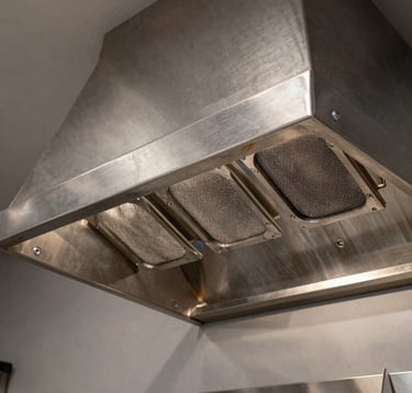 Close-up of a professional industrial kitchen hood in a high-end Brazilian restaurant. The stainless steel has a refined brushed finish, showing precision welding and high-performance inertial filters. Sophisticated technical lighting.