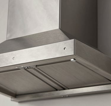 Detailed architectural close-up of the precision-engineered stainless steel surface of a range hood. The texture is brushed inox, showing clean lines and sophisticated metallic reflections under soft studio lighting.