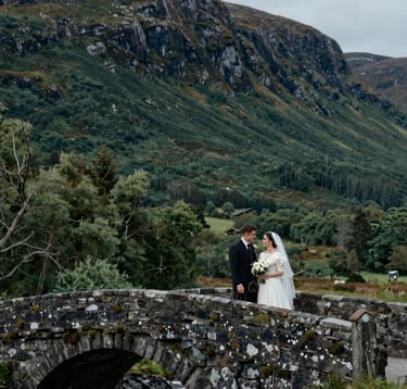 A wide cinematic shot of a couple standing on a stone bridge in the Scottish Highlands. The landscape is a mix of dark forest green and deep slate tones. The atmosphere is evocative of a traditional Northern European / Scottish wedding film.