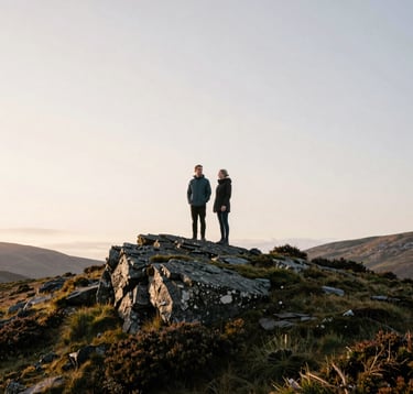 A Northern European / Scottish couple standing on a rugged hilltop at sunrise. The sky is a soft off-white. They are surrounded by sage green heather and dark slate rocks. The composition is expansive, showcasing the scale of the wild Scottish landscape.