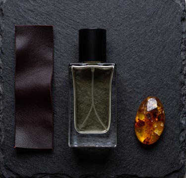 A top-down, minimalist flat lay of fragrance-related objects: a heavy glass bottle, a strip of dark leather, and a piece of raw amber on an obsidian black slate surface. Precise, high-end editorial style.