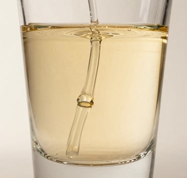 Macro photography of golden-hued perfume liquid diffusing through clear water in a crystal glass. The lighting is crisp and editorial, highlighting the fluid textures against a bone white background.