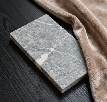 A macro photograph of high-end finishing materials. A slab of muted stone grey marble with white veining rests against a piece of dark ebony textured wood. A warm sand taupe velvet swatch is draped nearby. Clean, bright, and luxurious lighting.