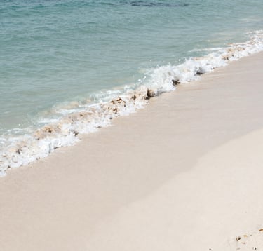 High-angle photography of turquoise ocean waves gently washing over fine white sand on a secluded Brazilian beach. The lighting is bright and clean, capturing the natural beauty of the South American coastline. Colors are dominated by muted blue and off-white.