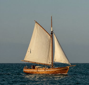 A serene photograph of a classic wooden sailboat gliding through deep teal waters off the North American coast. The sky is a muted gray-blue, and the sails are a soft warm white.