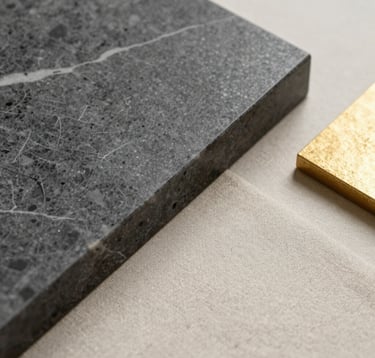 Macro photography of high-end interior materials: a piece of charcoal gray marble, a swatch of warm cream-beige linen, and a soft gold metallic accent piece arranged on a clean surface. Soft side lighting. International / High-end.