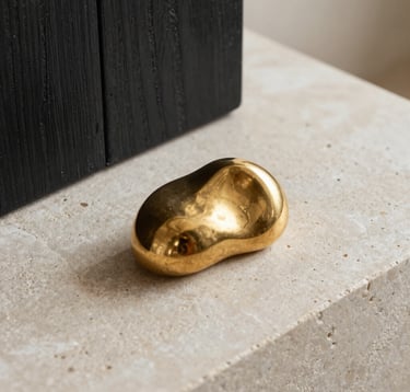 A macro photography shot of luxury interior details: a gold-toned sculptural object resting on a textured off-white stone surface next to a piece of dark charcoal oak. Soft, warm natural lighting. International / High-end.