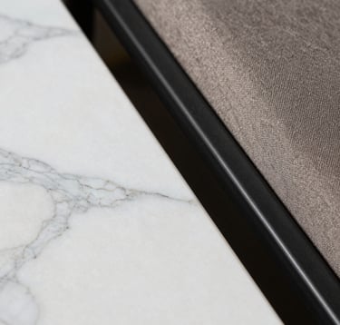 Close-up macro photography of high-end interior textures. A smooth slab of white Indian marble with delicate grey veins sits adjacent to a textured taupe fabric and a matte black metal frame. The lighting is soft and focused, emphasizing the premium quality of the materials.