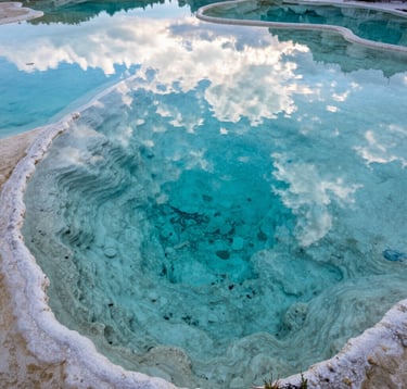 A close-up photograph of the turquoise mineral pools at the top of Hierve el Agua. The water is perfectly still, reflecting the soft sky blue of the Oaxacan clouds. Deep teal minerals are visible on the floor of the natural basin, surrounded by off-white calcified edges.