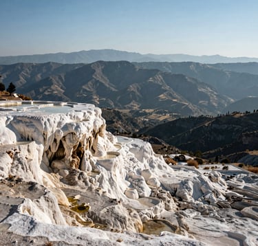 A landscape photograph showing the expansive view from the top of Hierve el Agua. The scene captures the layered mountain ranges of Oaxaca, Mexico, fading into a hazy soft blue distance. The foreground shows the textured mineral surface of the formations in off-white and charcoal tones.
